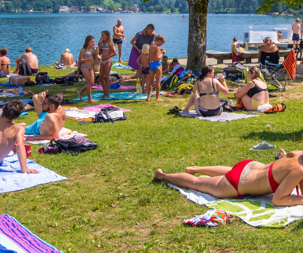 Tłum ludzi odpoczywających i opalających się na trawie nad jeziorem w słoneczny dzień, niektórzy w strojach kąpielowych, inni na kocach, symbolizuje spędzanie wolnego czasu. Artykuł na naszym portalu omawia kontrowersje związane z barierą językową na plażach.