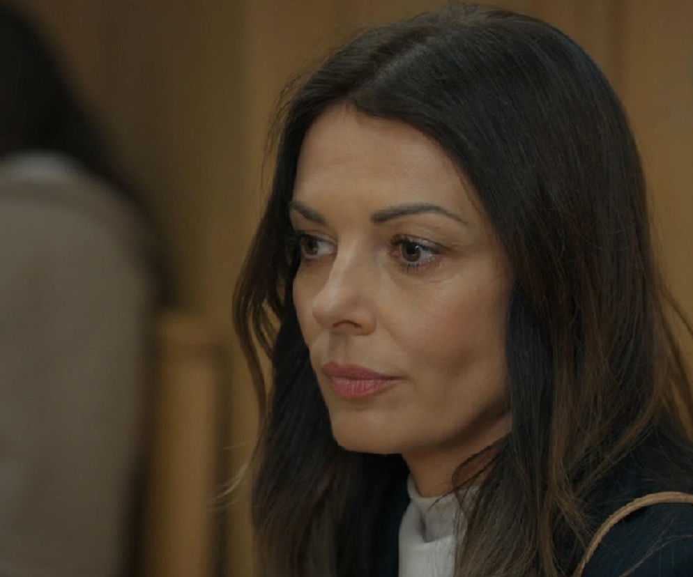 Kasia (Katarzyna Glinka) z serialu Barwy szczęścia z poważną miną podczas rozprawy sądowej, na zdjęciu zbliżenie na jej twarz i długie, brązowe włosy. Aktorka nosi ciemną marynarkę w prążki i biały golf, w tle rozmazana postać. Więcej o losach Kasi na Super Seriale.