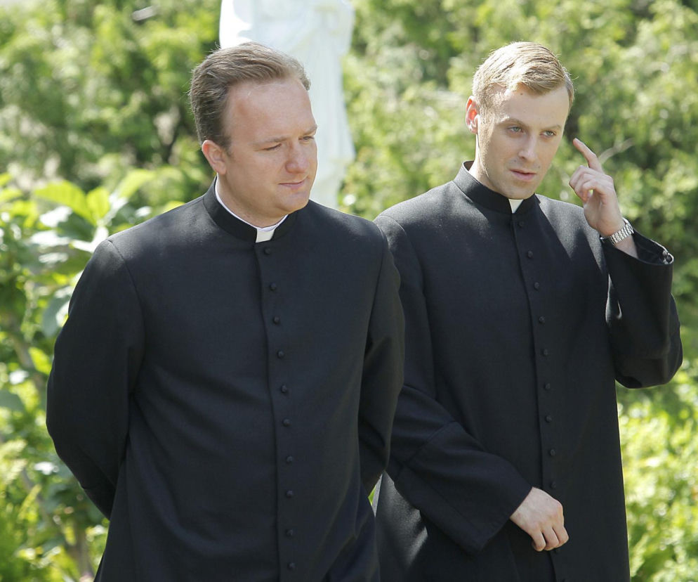 Proboszcz Robert (Bartłomiej Kasprzykowski) i wikary Maciej (Mateusz Rusin) z serialu Ranczo w czarnych sutannach, na tle zielonej roślinności, dyskutują. Księża z Wilkowyj znów zmienią się w detektywów, a ich kryminalne zagadki i nowa plebania znajdą się na Super Seriale.