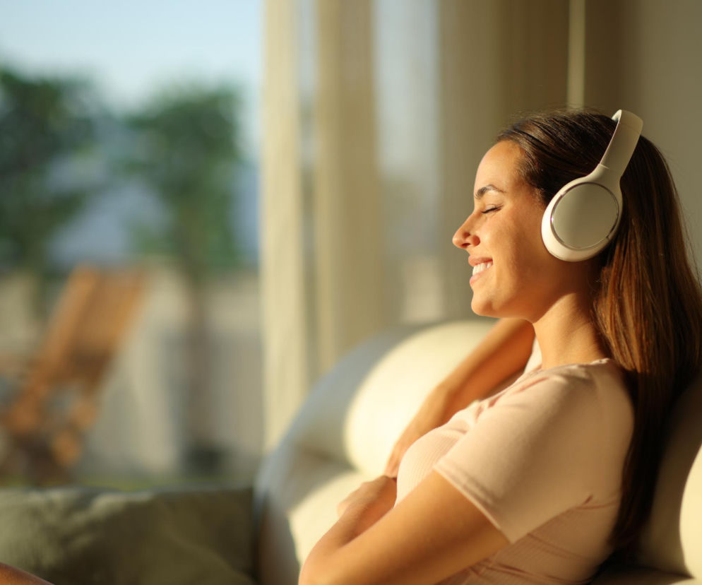 Uśmiechnięta kobieta z zamkniętymi oczami relaksuje się na kanapie, słuchając muzyki przez białe słuchawki, podczas gdy promienie słońca padają na jej twarz. Czytaj o sposobach na pozbycie się robaka słuchowego na Poradnik Zdrowie.