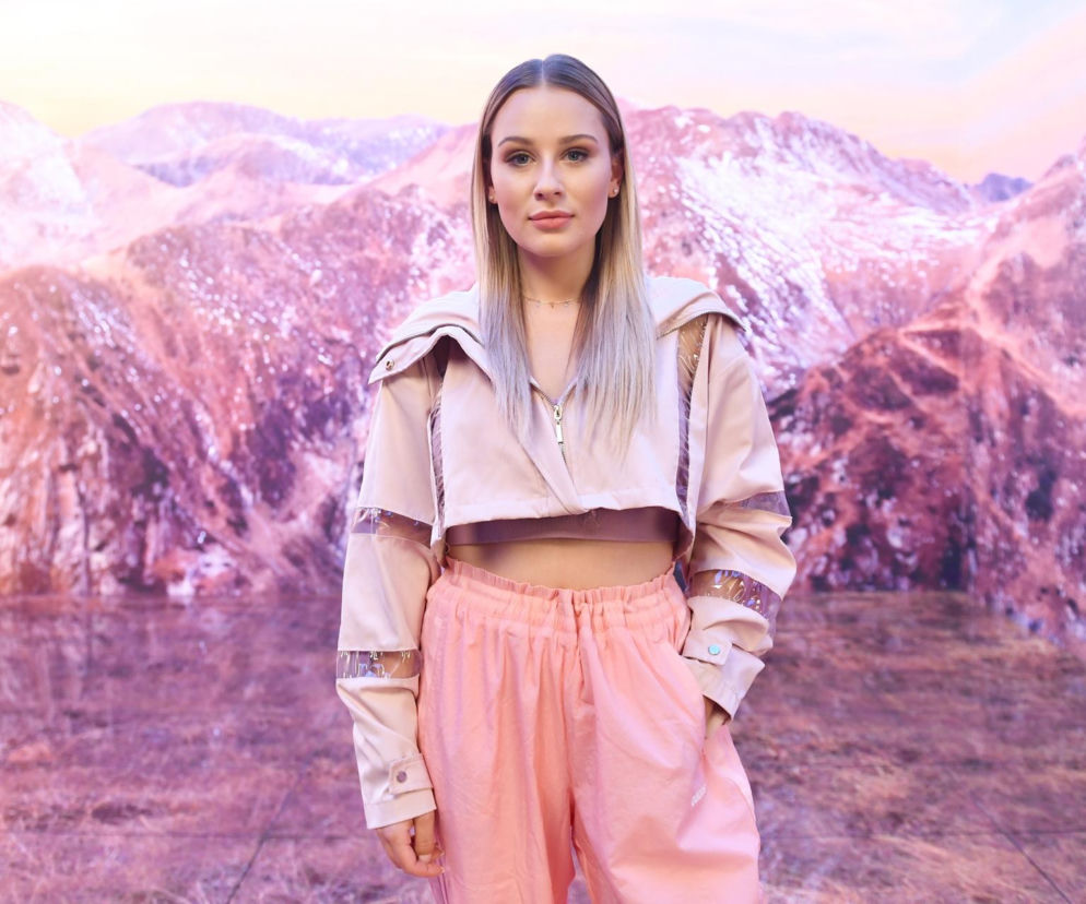 Influencerka Andziaks w różowym stroju sportowym na tle grafiki z górami. O podatkach jej firmy czytaj na SE Superbiz.