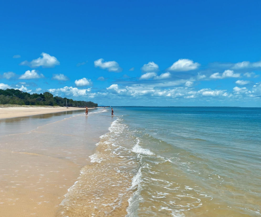Słoneczna plaża w Mielnie z delikatnymi falami obmywającymi brzeg i dwiema osobami w oddali, symbolizująca idealne warunki na weekendowy wypoczynek, o którym możesz przeczytać na naszym portalu.