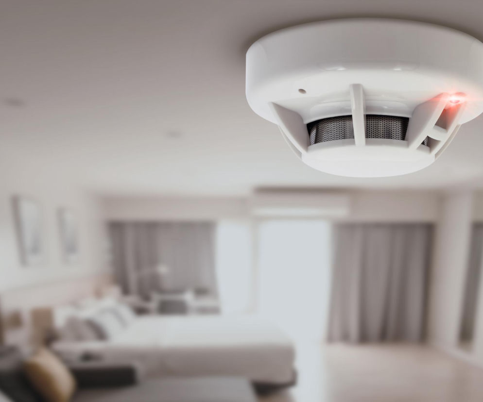 Biała czujka dymu z czerwoną diodą alarmową zamontowana na suficie nowoczesnego, jasnego pokoju. Obowiązek posiadania czujek dymu i czadu od 2030 roku w Polsce to temat, o którym przeczytasz więcej na Super Biznes.