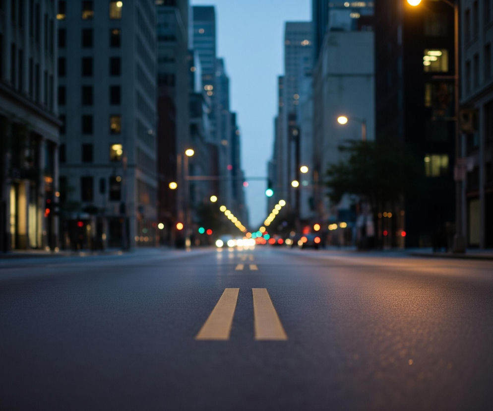 Asfaltowa ulica w centrum miasta rozciąga się w głąb kadru, z dwoma żółtymi liniami oddzielającymi pasy ruchu na pierwszym planie. Po obu stronach ulicy wznoszą się wysokie, szare budynki z licznymi oknami, niektóre z nich świecą ciepłym, żółtym światłem. W tle widoczne są rozmyte światła drogowe w kolorach czerwonym, zielonym i żółtym, oraz białe reflektory nadjeżdżających samochodów, co sugeruje wczesny wieczór lub świt.