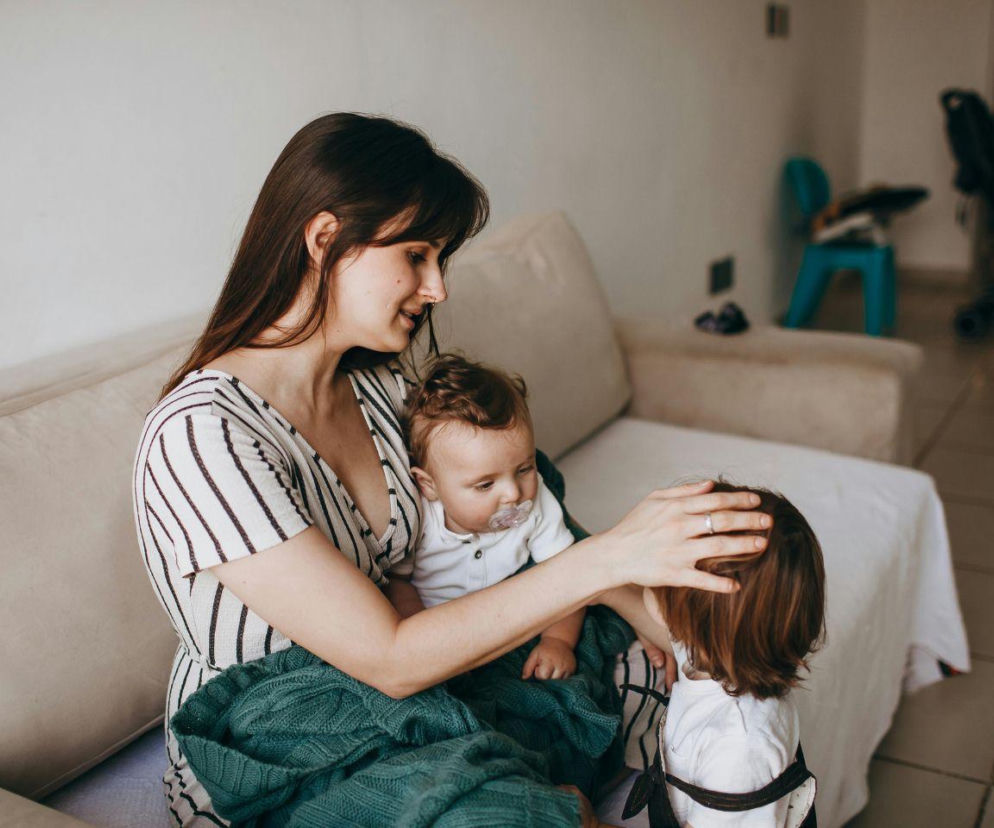 Mama siedzi na kanapie z niemowlakiem i głaszcze starsze dziecko po głowie. O wsparciu rodzeństwa przeczytasz na SE.