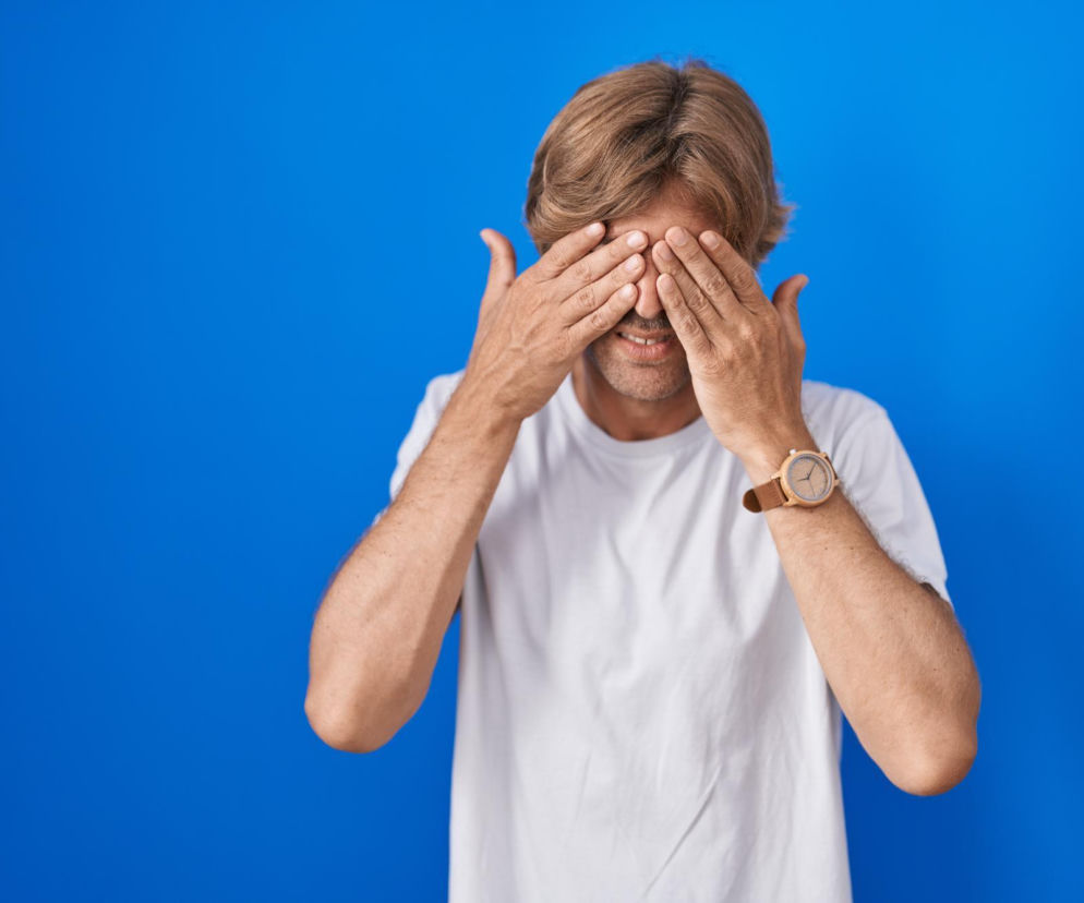 Mężczyzna z jasnymi włosami w białej koszulce na tle niebieskiej ściany zakrywa oczy dłońmi, symbolizując widzenie błysków po pocieraniu oczu. Artykuł na Poradnik Zdrowie wyjaśnia, kiedy błyski w oczach są niepokojące i mogą świadczyć o problemach ze wzrokiem.