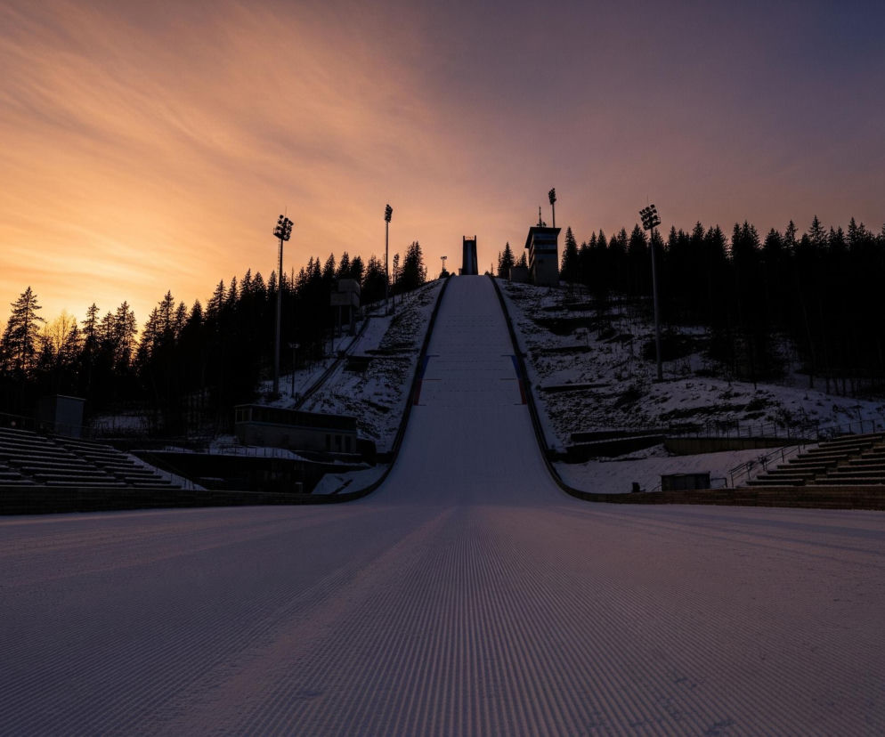 Ośrodek sportów zimowych oświetlony zachodzącym słońcem, dominuje skocznia narciarska pokryta białym, puszystym śniegiem z wyraźnymi, równoległymi śladami ratraka. Po obu stronach skoczni widoczne są trybuny z ławkami, a także wysokie, metalowe maszty z reflektorami. Horyzont wypełnia ciemny las i wzgórza, z których wyłaniają się sylwetki budynków i platform. Niebo, od koloru pomarańczowego na horyzoncie, przechodzi w fioletowy i błękitny w górnych partiach.