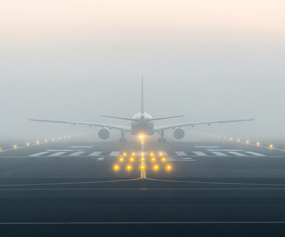 Samolot szerokokadłubowy stoi na pasie startowym, zwrócony tyłem do obserwatora, w warunkach gęstej mgły lub oparów. Jego ogon i skrzydła są wyraźnie widoczne, a silniki częściowo zasłonięte przez skrzydła. Pas startowy o ciemnoszarym kolorze jest oświetlony rzędami jaskrawopomarańczowych świateł, prowadzących w kierunku samolotu i dalej, w otaczającą mgłę. Na asfalcie widoczne są białe linie i oznaczenia, a horyzont rozjaśnia się delikatnym gradientem od bladoniebieskiego do jasnopomarańczowego, zlewającego się z mgłą.