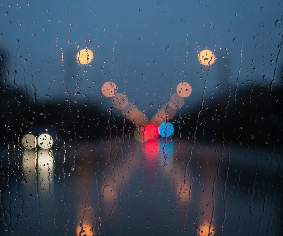 Przednia szyba z widocznymi licznymi kroplami deszczu i smugami wody dominuje obraz. Za nią rozmywają się jasne, rozproszone światła miejskie w odcieniach żółci, pomarańczy, czerwieni i błękitu, tworząc efekt bokeh. W centrum kadru widać dwa wyraźne, rozmyte światła: jedno czerwone po lewej i jedno niebieskie po prawej. Tło jest ciemnoniebieskie, przechodzące w czerń u dołu i po bokach, co sugeruje noc lub zmierzch.