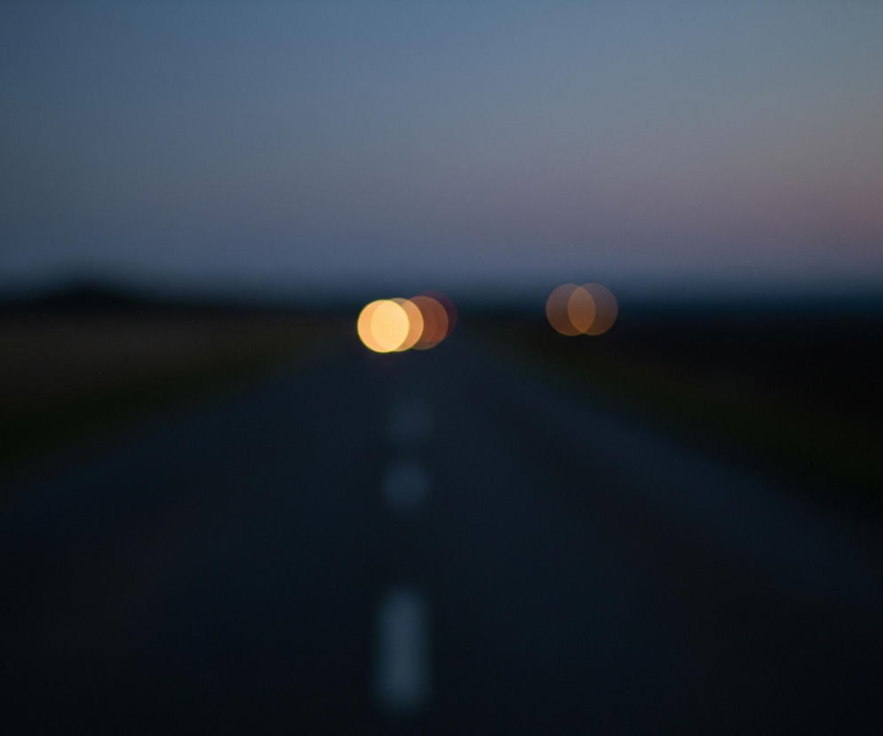 Prosta, ciemna droga asfaltowa rozciąga się w głąb obrazu, zajmując jego dolną połowę. Na drodze, na środku kadru, widoczne są rozmyte, białe paski, wyznaczające oś jezdni. W tle, na wysokości horyzontu, unoszą się dwie rozświetlone, okrągłe plamy w odcieniach pomarańczu i żółci, stanowiące prawdopodobnie światła pojazdu. Nad horyzontem ciemne niebo przechodzi od głębokiego granatu u góry, przez błękit, aż do jaśniejszych, lekko różowawych tonów tuż nad linią horyzontu.