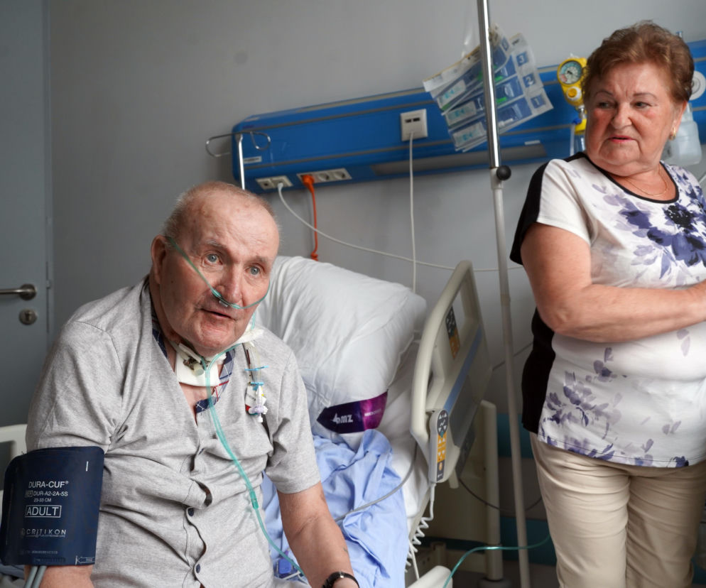 Pacjent Henryk Pietrowski z założoną kaniulą nosową i kołnierzem ortopedycznym, z mankietem do mierzenia ciśnienia na ramieniu, siedzi na łóżku szpitalnym, obok niego stoi jego żona, Marianna, opiekuje się nim. Ich historię o walce z chorobą i szczęśliwym końcu przeczytasz na portalu Poradnik Zdrowie.
