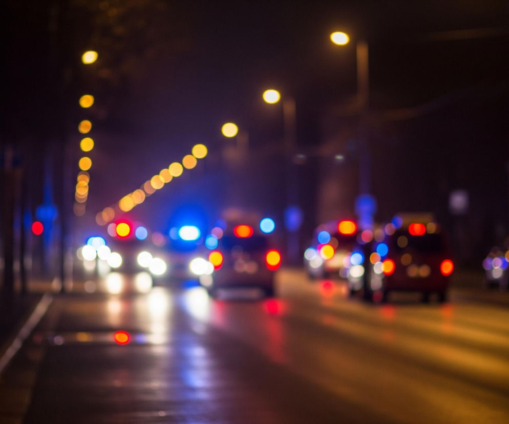 Rozmyty obraz przedstawia nocną ulicę miejską z widocznym ruchem pojazdów. W centrum dominują jasne, rozproszone światła samochodów, w tym białe reflektory, czerwone światła tylne oraz wyraźne niebieskie sygnały świetlne, sugerujące obecność pojazdów uprzywilejowanych. Wzdłuż ulicy po lewej stronie znajdują się drzewa, a po prawej budynki, z których widać rozproszone żółte światła latarni ulicznych w górnej części kadru. Całość utrzymana jest w ciemnej tonacji z dominującymi kolorami świateł: żółtym, czerwonym, niebieskim i białym.