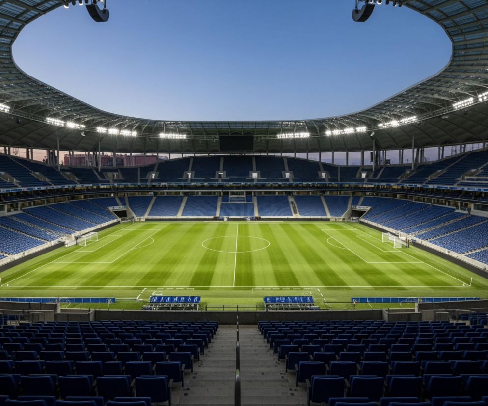 Rozległy, nowoczesny stadion piłkarski, widziany z trybun, jest pusty, z wyjątkiem równo skoszonej, zielonej murawy rozświetlonej przez intensywne reflektory umieszczone pod przeszklonym dachem. Niebieskie siedzenia trybun, rozciągające się w wielu rzędach wokół boiska, są całkowicie puste, a nad nimi rozciąga się metalowa konstrukcja dachu z widocznymi, jasnymi światłami. Jasne, niebieskie niebo widoczne jest ponad otwartym centralnym fragmentem dachu.