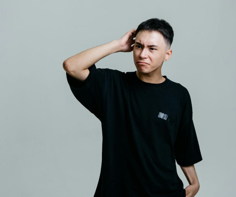 Młody mężczyzna w czarnej koszulce drapie się po głowie z wyraźnym grymasem zaniepokojenia i dyskomfortu. Jego zaciśnięte usta i zmarszczone brwi podkreślają problem swędzącej skóry głowy i przetłuszczających się włosów, o którym możesz przeczytać na portalu Poradnik Zdrowie.