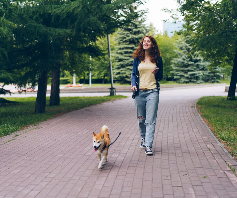 Uśmiechnięta kobieta spaceruje z psem rasy shiba inu w parku. O miejscach dla psów w Poznaniu przeczytasz na Eska Poznań.