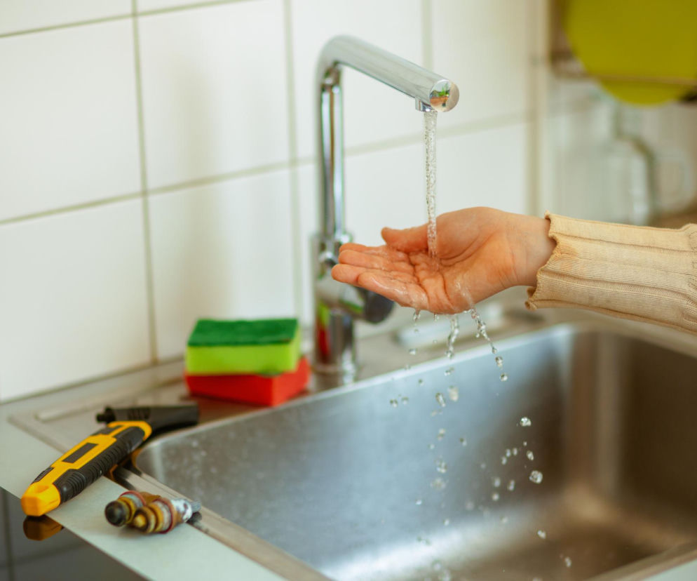 Ręka osoby pod strumieniem chłodnej wody z kranu w kuchni, co symbolizuje pierwszą pomoc przy oparzeniu, o czym więcej przeczytasz na Poradnik Zdrowie. Ujęcie skupia się na bieżącej wodzie i dłoni w jasnym, domowym wnętrzu.
