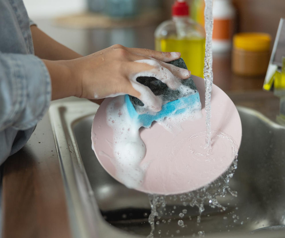 Ręce myjące różowy talerz gąbką z niebieską warstwą ścierną i dużą ilością piany pod strumieniem wody w zlewie, co nawiązuje do tematu artykułu o higienie gąbek do naczyń. Dowiedz się więcej na Poradnik Zdrowie.