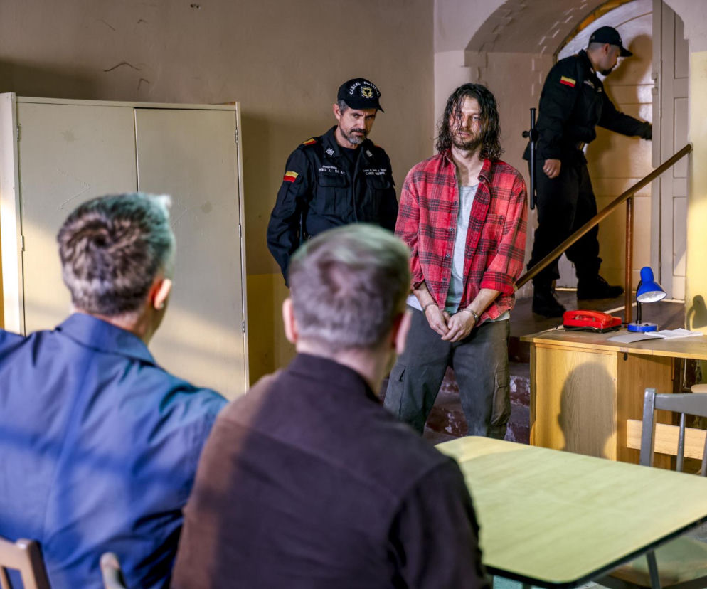 Mateusz Kościukiewicz jako Paweł Krzyżanowski w kajdankach w więzieniu w Kolumbii, pod nadzorem strażników, spogląda na Janka i Artura, siedzących przy stole. Scena zapowiada dramatyczny powrót bohatera do Pierwszej miłości, o czym więcej na Super Seriale.