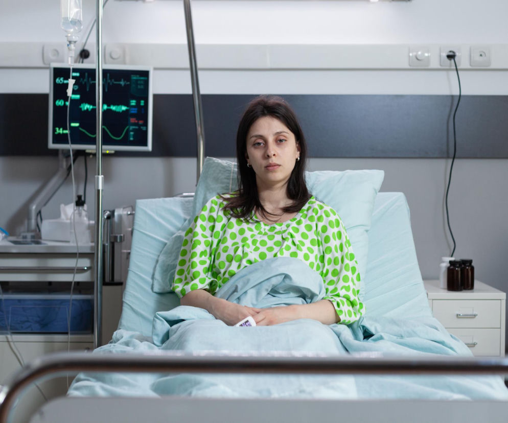 Pacjentka z miastenią leży w szpitalnym łóżku, trzymając pulsoksymetr. Wygląda na zmęczoną, obok niej monitor z parametrami życiowymi oraz kroplówka. Na portalu Poradnik Zdrowie znajdziesz informacje o nowoczesnym leczeniu miastenii.