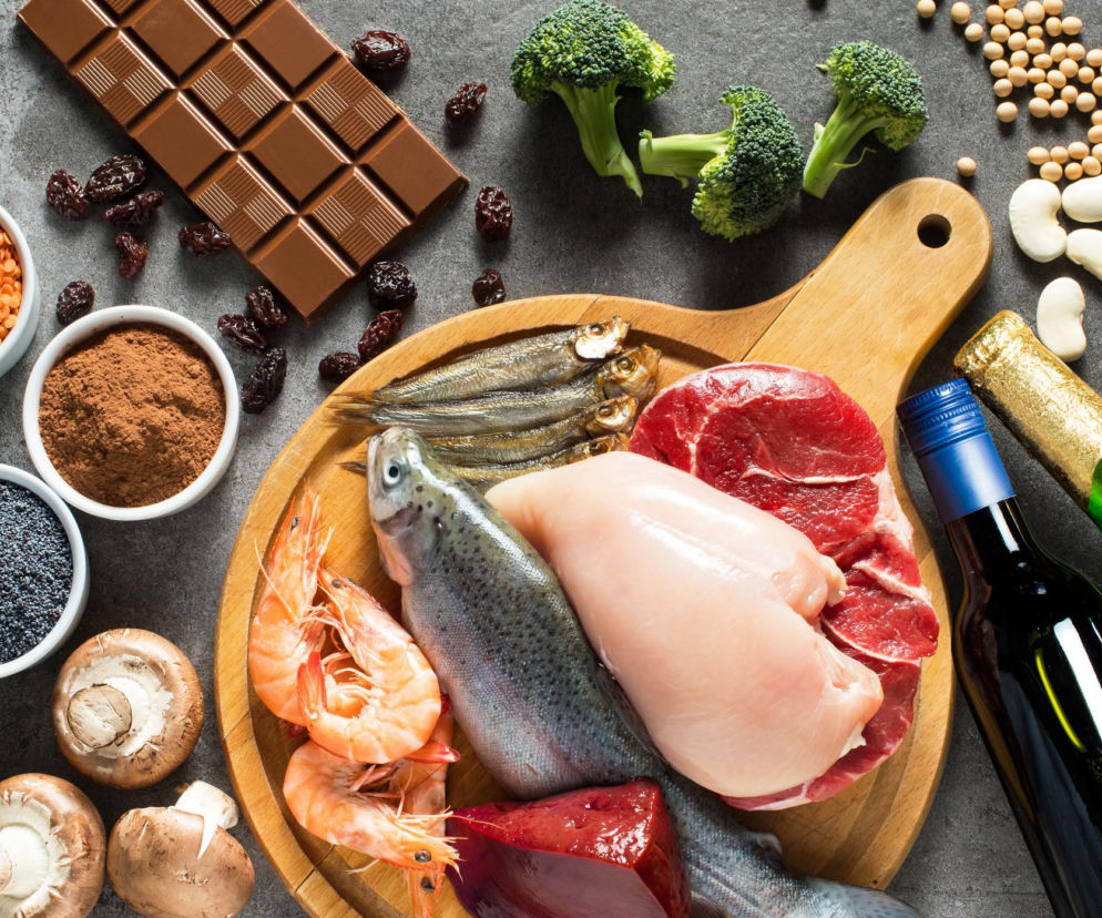 Kompozycja produktów spożywczych ułożonych na ciemnej, kamiennej powierzchni. Na drewnianej desce świeże mięso (filet z kurczaka, kawałek wołowiny, ryba pstrąg, małe rybki, krewetki). Obok: czekolada, brokuły, suszone śliwki, soczewica, kakao, mak, pieczarki, fasola, soja oraz butelki wina i szampana. Poradnik Zdrowie przypomina, że odpowiednie jedzenie poprawia nastrój.