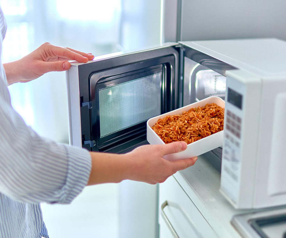 Osoba w paski koszuli wkłada prostokątny pojemnik z ciepłym posiłkiem do mikrofalówki, co symbolizuje bezpieczne i szybkie podgrzewanie jedzenia. Ten obraz ilustruje temat bezpiecznego korzystania z mikrofalówek, o czym więcej można przeczytać na portalu Poradnik Zdrowie.