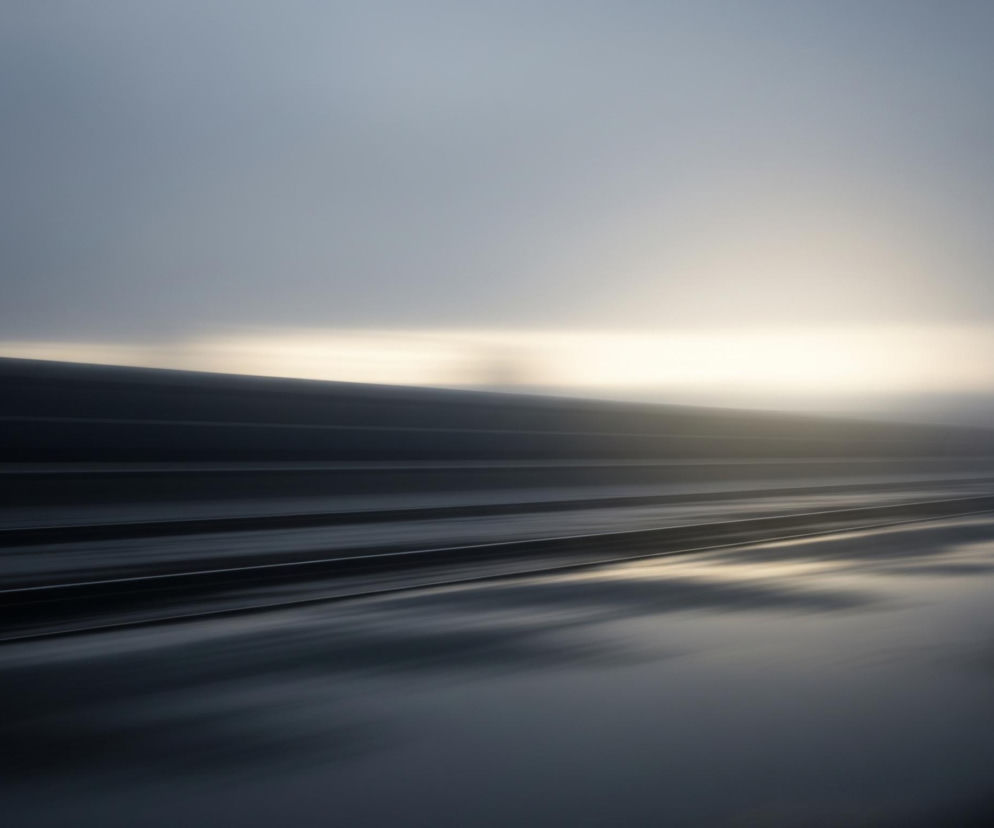 Ciemną, abstrakcyjną kompozycję z rozmytymi poziomymi liniami. Górna część obrazu to jednolite, szare niebo, przechodzące w jaśniejszy, mleczny odcień bieli ku prawej stronie horyzontu. W dolnej części widoczne są szerokie, poziome pasy w ciemnych odcieniach szarości i czerni, sprawiające wrażenie falistego terenu lub torów, z refleksami jaśniejszego światła odbijającego się na ich powierzchni. Rozmycie i wydłużone smugi światła sugerują ruch lub długą ekspozycję.
