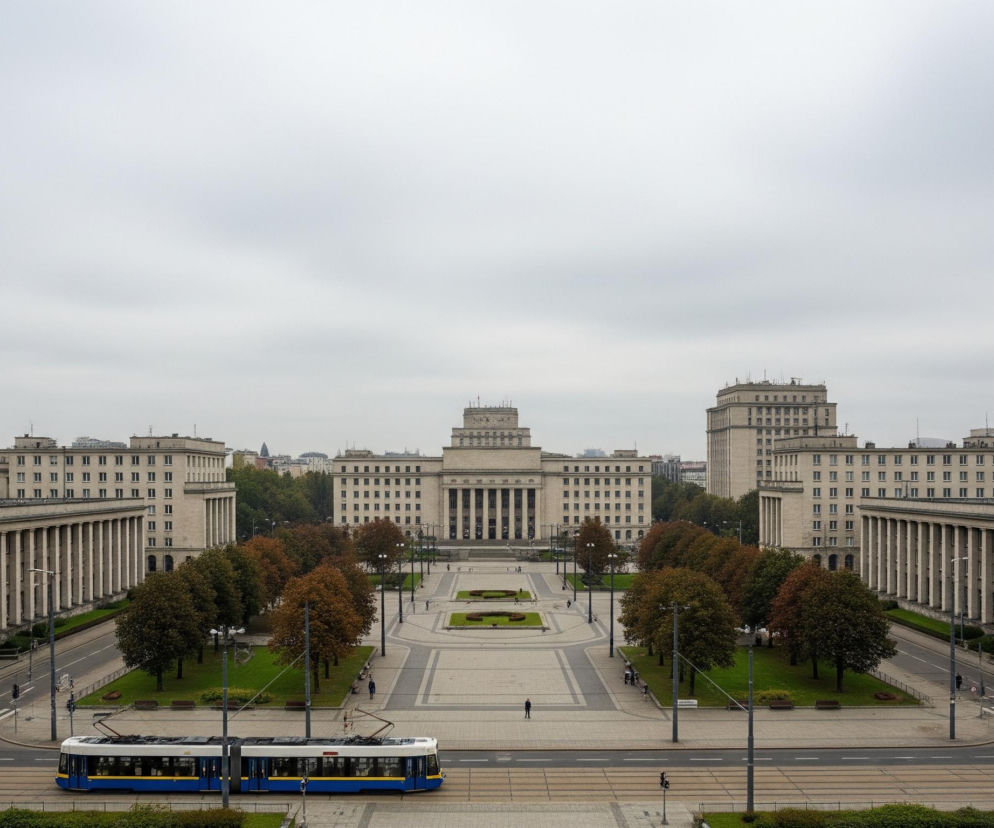 Szeroki widok lotniczy przedstawia duży, symetryczny plac miejski otoczony monumentalnymi budynkami w odcieniach szarości i beżu pod pochmurnym, jasnym niebem. W centrum kompozycji znajduje się długi, prostokątny pas zieleni z rzędem drzew o jesiennych barwach, oddzielający dwie jezdnie z torami tramwajowymi. Na pierwszym planie po lewej stronie widać niebiesko-biały tramwaj jadący w prawo, natomiast dalej na placu widoczne są pojedyncze postacie ludzkie. W tle dominuje wysoki budynek z kolumnami i centralną wieżą, a po jego bokach rozciągają się kolejne, podobne architektonicznie konstrukcje.