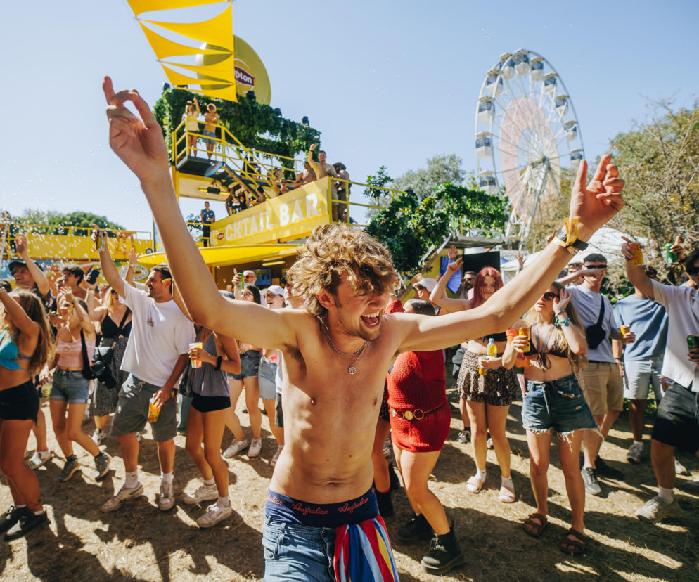 Mężczyzna bez koszulki tańczy z uniesionymi rękami na Sziget Festival. W tle diabelski młyn i bar. Czytaj na Eska.