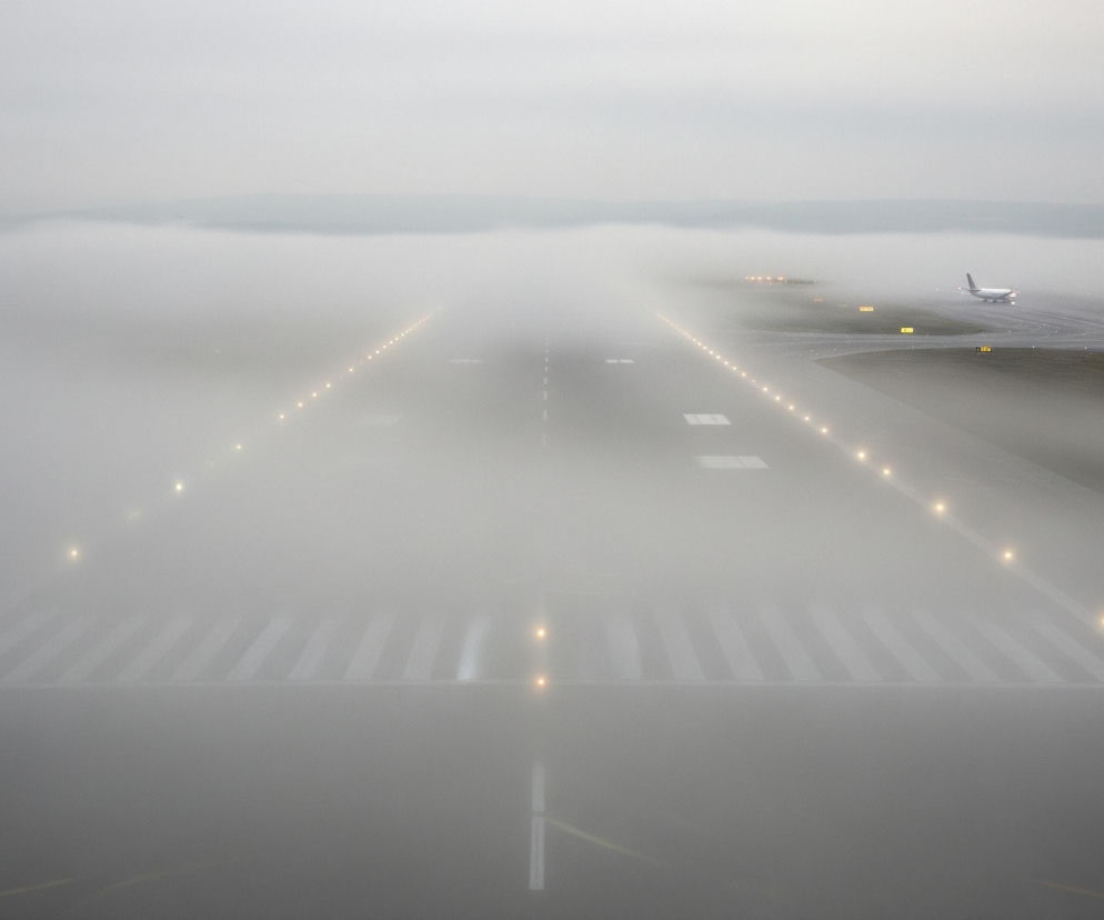 Płyta lotniska w szarej mgle rozciąga się w głąb obrazu, z pasem startowym wyraźnie widocznym pośrodku. Białe, prostokątne oznaczenia malowane są na ciemniejszej nawierzchni pasa, otoczonego przez rzędy intensywnie świecących, żółtych świateł, rozchodzących się w perspektywie. W prawej części obrazu, na skrzyżowaniu dróg kołowania, stoi biały samolot z fioletowymi elementami na ogonie i skrzydłach, którego światła nawigacyjne są zapalone. Całość otoczona jest gęstą mgłą, która zasłania horyzont, nadając scenie eteryczny i tajemniczy wygląd.