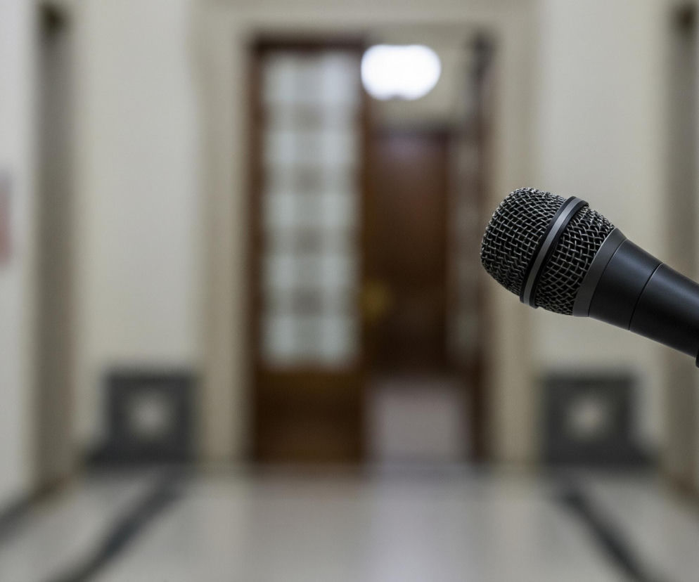 Czarny mikrofon z metalową, siatkową główką znajduje się na pierwszym planie po prawej stronie. Mikrofon jest podłączony do ciemnego statywu mikrofonowego, widocznego w dolnym prawym rogu. Tło jest rozmyte i przedstawia wnętrze budynku z beżowymi ścianami oraz ciemnymi drzwiami z przeszkleniami w centrum. Podłoga składa się z beżowych i ciemnych, prawdopodobnie czarnych, płytek, tworzących wzór.