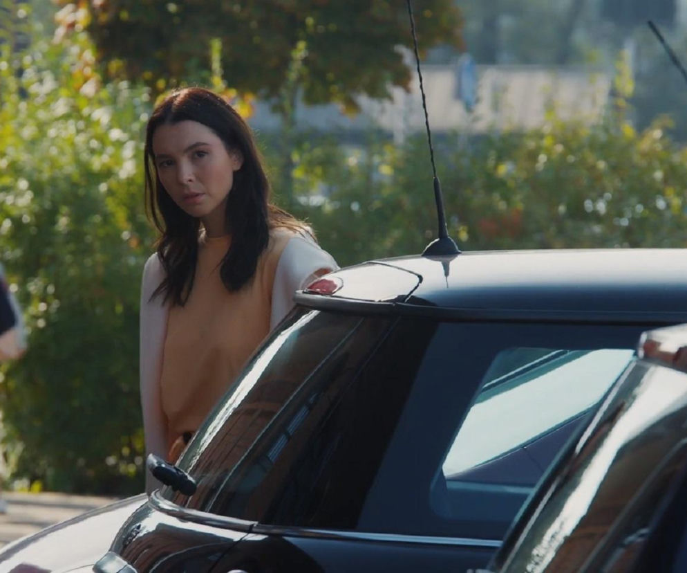 Aktorka Marcelina Kieres jako Paula, z poważną miną, w tle rozmazane sylwetki ludzi i zieleń, obok ciemny samochód. Opis sceny zemsty Pauli na Natalce z M jak miłość znajdziesz na Super Seriale.