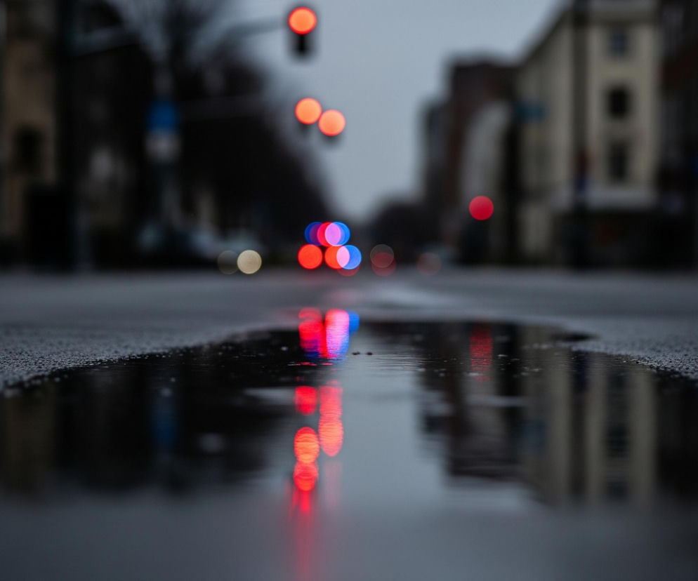 Głównym elementem jest kałuża na pierwszym planie, w której odbijają się rozmyte światła uliczne. Na wodzie widoczne są intensywne, czerwone i niebieskie refleksy świateł, przypominające owalne kształty. Ponad kałużą rozciąga się ciemna, mokra nawierzchnia drogi, na której widoczne są drobne kropelki wody.

W oddali, tło jest silnie rozmyte, ukazując kontury budynków w szarych i beżowych barwach oraz kolejne, jasne czerwone i pomarańczowe światła uliczne, również w formie okręgów. Niebo jest jednolicie szare, co wskazuje na pochmurny dzień. Ogólny nastrój obrazu jest spokojny i lekko melancholijny, z dominacją ciemnych, chłodnych barw.