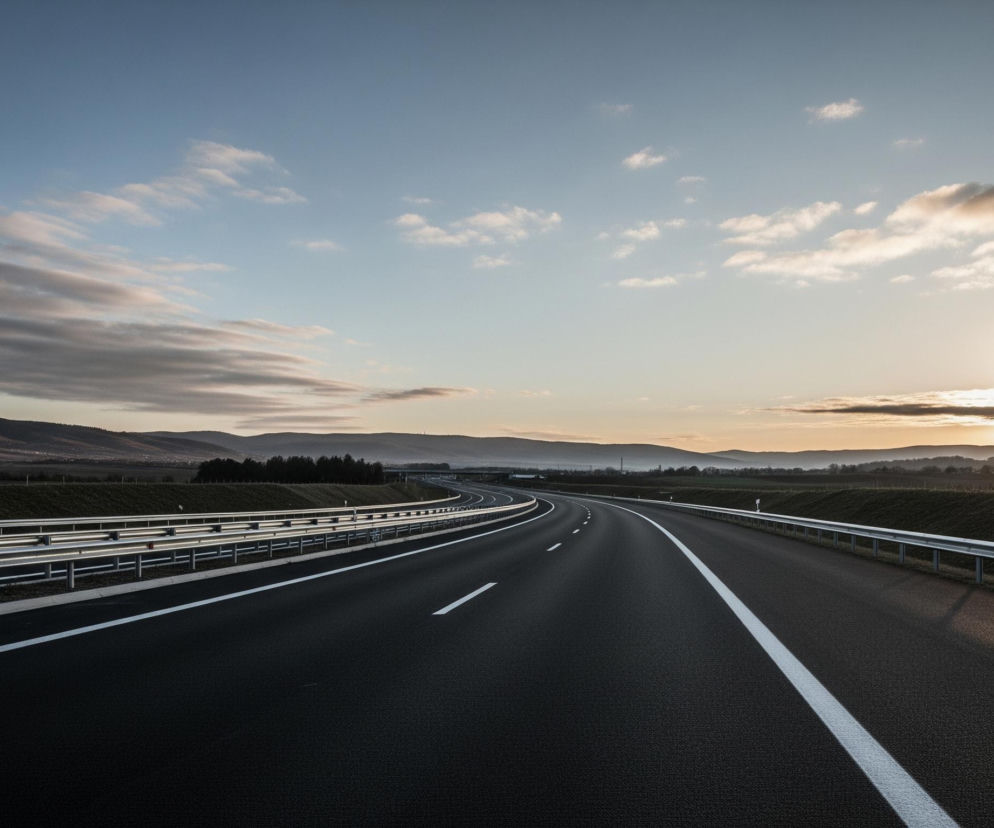 Asfaltowa autostrada z dwoma pasami ruchu widoczna jest na pierwszym planie, z białymi, ciągłymi i przerywanymi liniami rozdzielającymi pasy. Po lewej stronie jezdni znajduje się metalowa barierka ochronna, za którą rozciąga się zielony nasyp, a następnie pasmo drzew i pagórków, na horyzoncie zlewających się z szarofioletową mgiełką. Po prawej stronie drogi również ciągnie się barierka, za którą widoczny jest trawiasty teren i rozległe pola, a dalej zarys zabudowań. Niebo jest błękitno-szare, z licznymi chmurami w odcieniach bieli, szarości i pomarańczu, a słońce, świecące nisko nad horyzontem, widoczne jest po prawej stronie kadru, emitując ciepłe, złote światło.