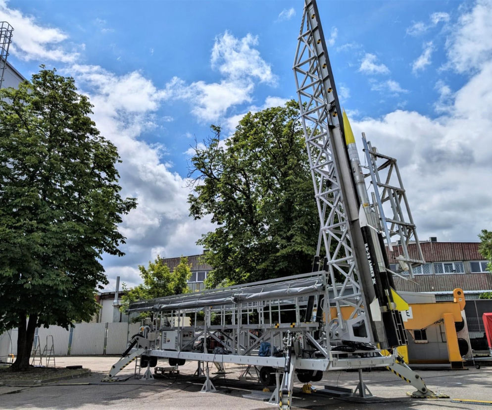 Polska rakieta suborbitalna ILR-33 Bursztyn 2K gotowa do testów na wyrzutni, z jasnożółtą osłoną stożkową, otoczona drzewami i budynkami w tle, co świadczy o postępach w programie kosmicznym, o którym można przeczytać na naszym portalu.