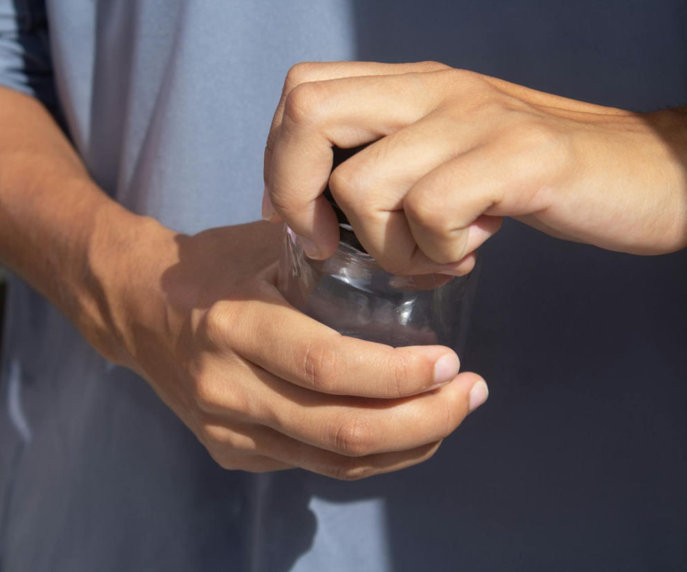 Osoba próbuje odkręcić wieczko słoika, co ilustruje problem ze słabym uściskiem dłoni i trudnościami w codziennych czynnościach. Na portalu Poradnik Zdrowie znajdziesz sposoby na wzmocnienie siły chwytu i zapobieganie bólom nadgarstków.