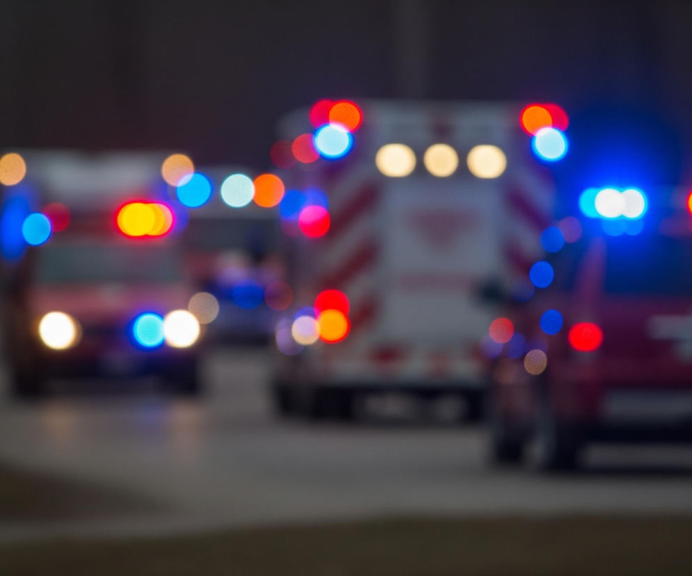 Rozmyte światła pojazdów ratunkowych wypełniają kadr, tworząc serię kolorowych okręgów w odcieniach czerwieni, błękitu i pomarańczu na ciemnym, niewyraźnym tle. Trzy pojazdy są widoczne w rzędzie, z których środkowy wydaje się być ambulansem z ukośnymi pasami na tyle, a dwa zewnętrzne to prawdopodobnie samochody osobowe. Świecące punkty światła, symulujące lampy błyskowe, dominują scenę, sugerując ruch i pilną sytuację.