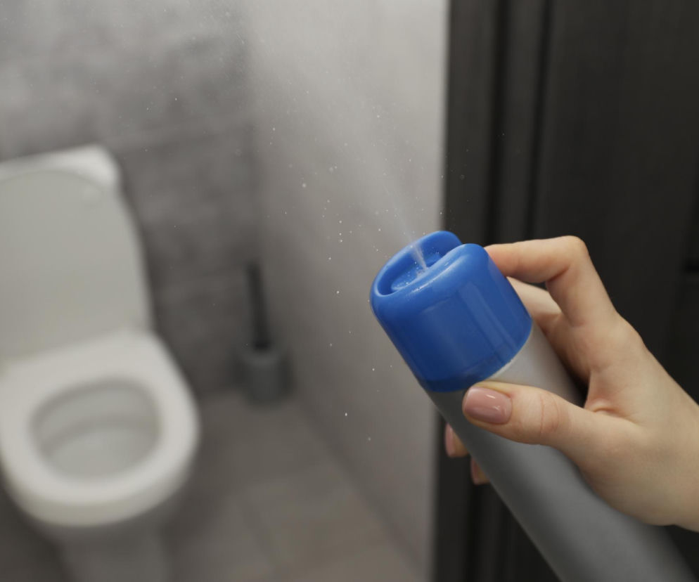 Dłoń trzymająca odświeżacz powietrza w aerozolu, z widoczną w tle niezamkniętą muszlą klozetową. Odświeżacz wydobywa mgiełkę. Ta fotografia ilustruje temat dbałości o higienę w łazience i rozprawia się z mitem o zamykaniu klapy sedesu, o czym przeczytasz na Poradnik Zdrowie.