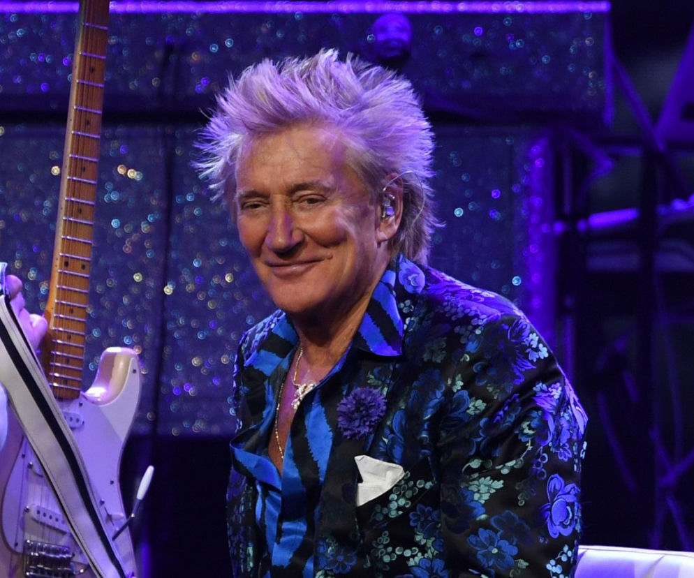 Uśmiechnięty Rod Stewart w niebieskiej marynarce na tle gitary. O problemach zdrowotnych gwiazdy przeczytasz na EskaRock.