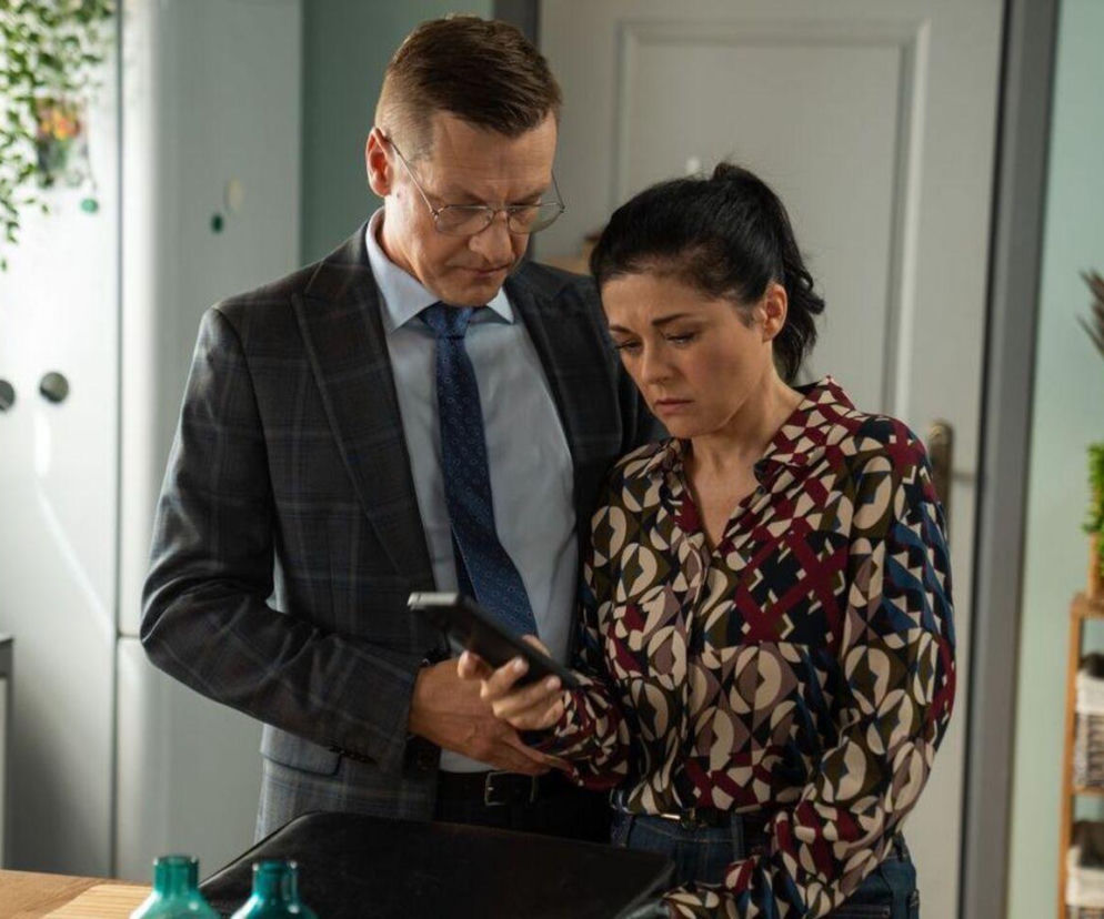 Piotrek Zduński (Marcin Mroczek) i Kinga Zduńska (Katarzyna Cichopek) ze „M jak miłość” w ich kuchni, razem pochylają się nad telefonem. Piotr w garniturze i krawacie, Kinga w wzorzystej bluzce. Na portalu Super Seriale czytaj o losach bohaterów.