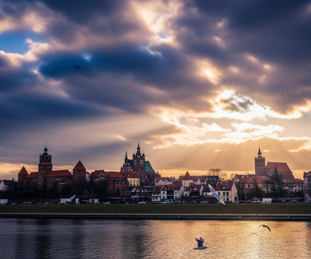 Szeroka panorama miasta ukazuje linię brzegową rzeki oraz zabudowania na wzgórzu, rozświetlone promieniami zachodzącego słońca, które przenikają przez rozległe, ciemne chmury. W lewej części kompozycji dominuje kompleks średniowiecznych budowli z cegły, składający się z zamkowych wież i zabudowań, przylegający do dużego kościoła z ostro zakończonymi wieżami i zielonym dachem. Po prawej stronie widać mniejsze budynki mieszkalne z czerwonymi dachami oraz dalsze wieże architektoniczne, a całość odbija się w złotawych, falujących wodach rzeki na pierwszym planie, gdzie trzy ptaki o jasnym upierzeniu lecą nisko nad powierzchnią.