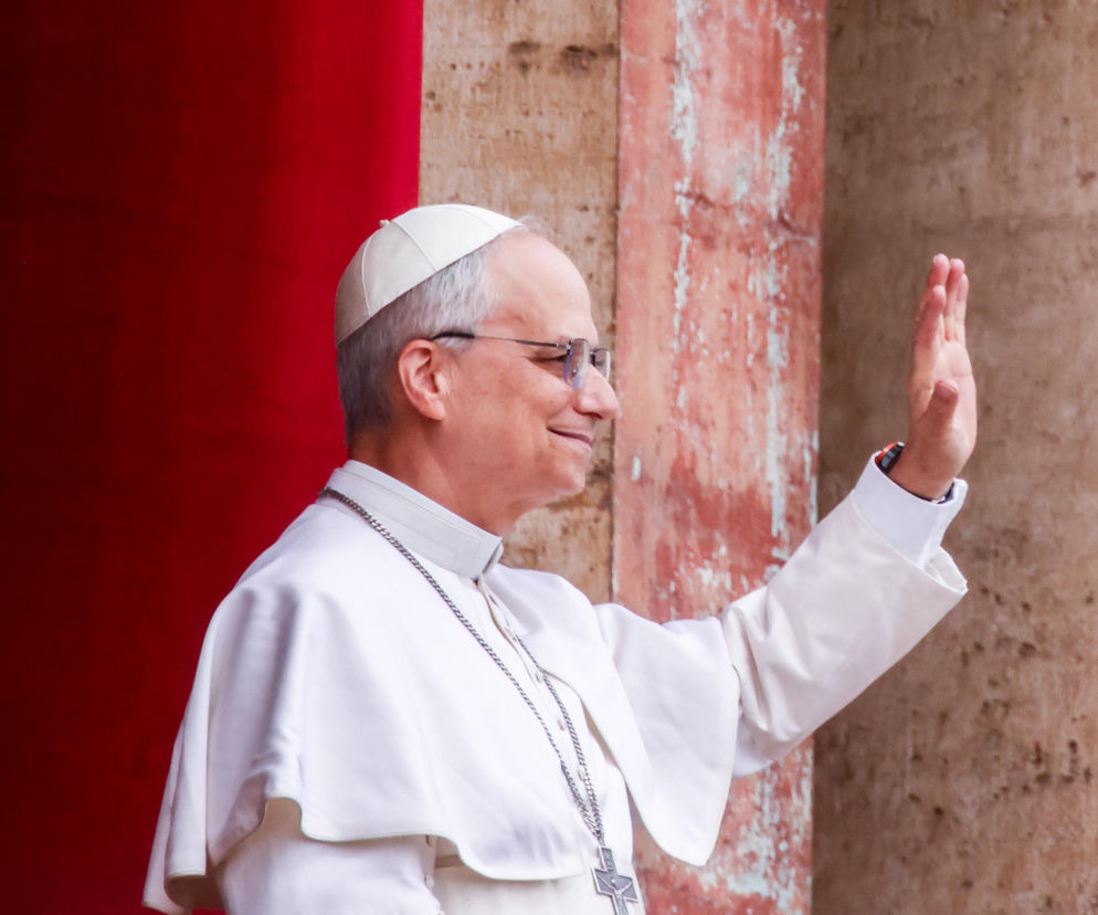 Papież Leon XIV w białej sutannie i okularach uśmiecha się i macha ręką. O kondolencjach dla rodzin przeczytasz w Eska.