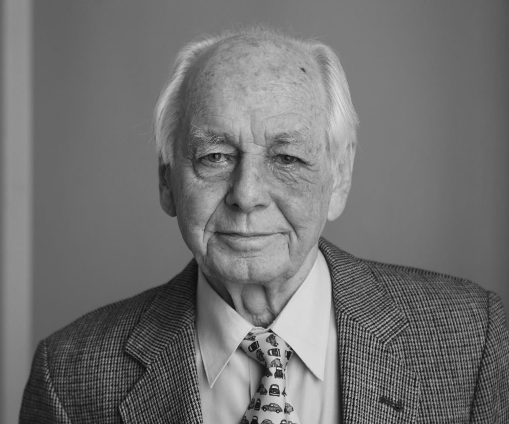Andrzej Martynkin, ceniony dziennikarz sportowy i sportowiec, w czarno-białym ujęciu portretowym, patrzący wprost na widza. Ubrany w elegancką marynarkę w kratę i koszulę z krawatem w motywy samochodowe. O jego bogatym życiu i karierze przeczytasz więcej na naszym portalu.