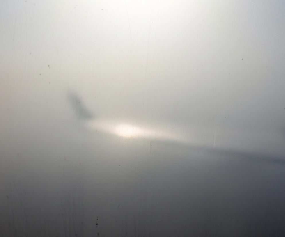 Rozmyty obraz z perspektywy wnętrza samolotu, spoglądając przez okno. Lewe skrzydło samolotu jest słabo widoczne na horyzoncie, zanurzone w gęstej, szarej mglistości lub chmurach. Jasne, pomarańczowo-białe światło, prawdopodobnie słońca, prześwituje z lewej strony obrazu, rzucając odblask na powierzchnię skrzydła. Całe ujęcie jest zamglone i pełne drobnych kropek, przypominających pył lub kropelki wody na szybie.