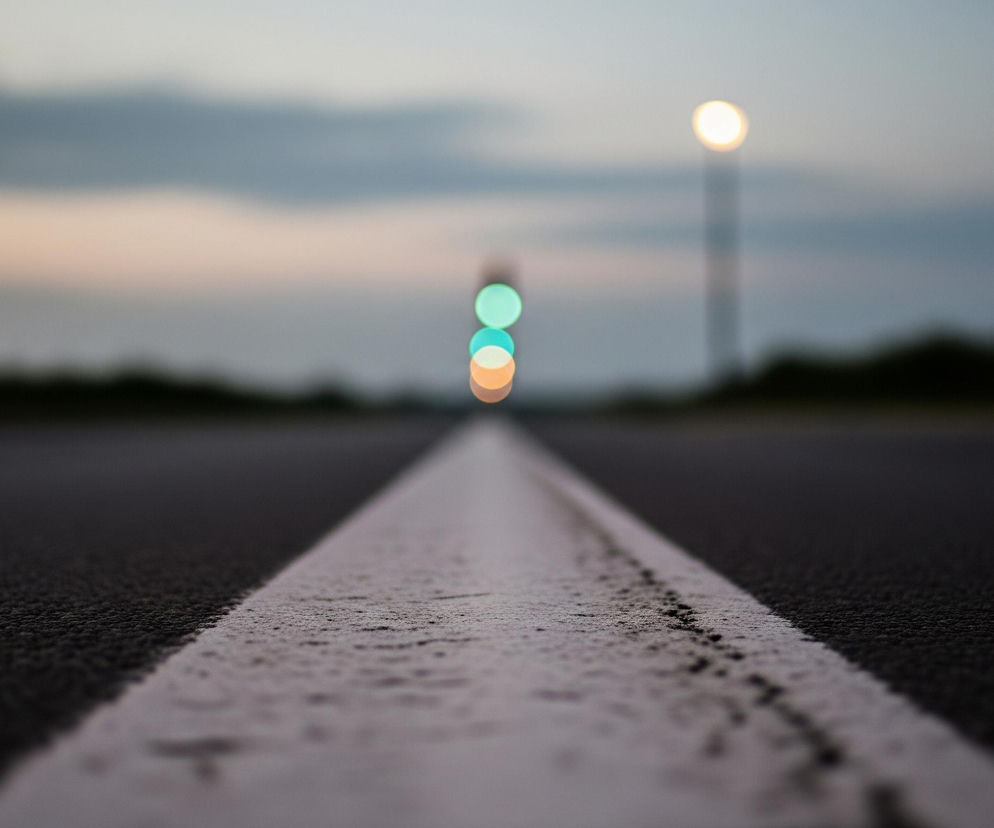 Ciemna, asfaltowa droga rozciąga się w głąb obrazu, a pośrodku niej biegnie biała, wąska linia, która staje się cieńsza i bledsza w oddali. Po prawej stronie drogi, w tle, widoczny jest długi, smukły słup z jasnym, rozmazanym punktem świetlnym na szczycie. Dalej, po lewej stronie tego słupa, pojawia się seria trzech okrągłych, rozmytych świateł: zielone na górze, turkusowe pośrodku i pomarańczowe na dole. Horyzont składa się z ciemnych, niewyraźnych sylwetek, prawdopodobnie drzew lub zabudowań, a nad nimi rozciąga się niebo o zmieniających się odcieniach szarości, błękitu i delikatnego różu, typowych dla świtu lub zmierzchu.
