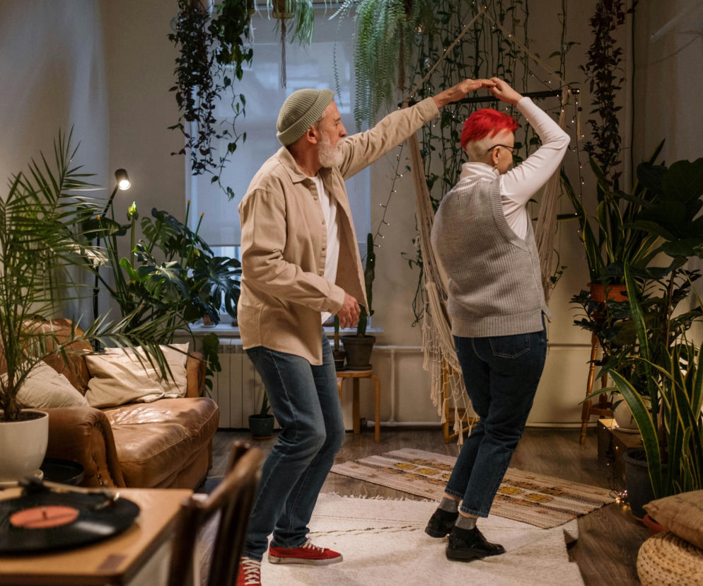 Starsza para tańczy w salonie, mężczyzna w beżowej koszuli i czapce, kobieta z krótkimi rudymi włosami w szarej kamizelce, otoczeni bujną roślinnością. Ta domowa aktywność to świetny sposób na poprawę kondycji i samopoczucia, o czym więcej przeczytasz na Poradnik Zdrowie.