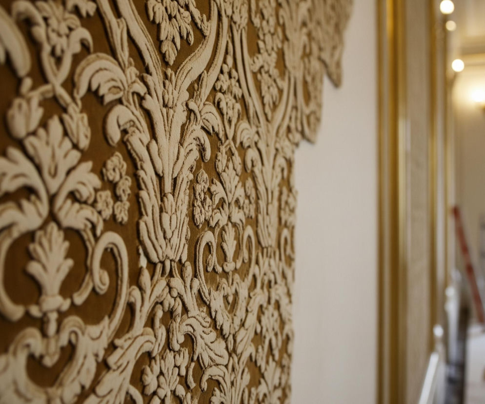 Na pierwszym planie, z lewej strony, widoczna jest brązowa ściana pokryta beżowymi, reliefowymi zdobieniami w stylu barokowym, składającymi się z motywów roślinnych, liści i kwiatów. Zdobienia są lekko postrzępione na brzegach i wydają się być wykonane z delikatnego materiału. Po prawej stronie tej ściany znajduje się fragment gładkiej, kremowej ściany, a za nią, w głębi, widać rozmazane tło przedstawiające wnętrze pomieszczenia z oknem, zasłonami i elementami konstrukcyjnymi, być może rusztowaniem, oraz punktowe źródła światła na suficie i kinkiet na ścianie.