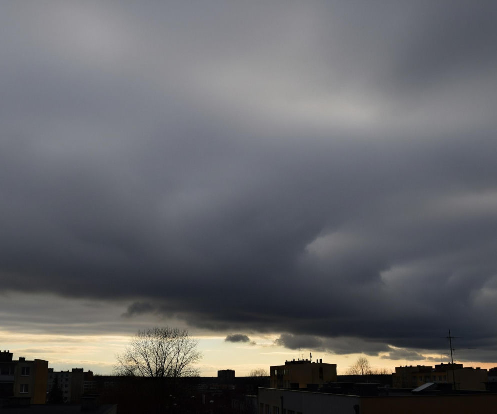 Górną część obrazu zajmują ciężkie, ciemnoszare chmury o zróżnicowanej fakturze, z jaśniejszymi fragmentami na górze i po prawej stronie. Pod nimi rozciąga się wąski pas jaśniejszego, żółtawo-pomarańczowego nieba, widocznego tuż nad horyzontem. Na dole widać sylwetki budynków mieszkalnych o płaskich dachach, z antenami i innymi konstrukcjami, oraz pozbawione liści drzewa, wszystkie ciemne i niewyraźne, odcinające się na tle nieba. Scena przedstawia prawdopodobnie późne popołudnie lub wieczór, z dominującą, ponurą atmosferą.