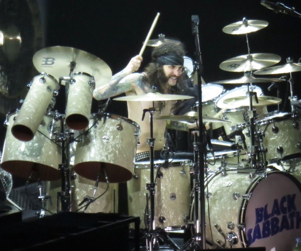 Perkusista Tommy Clufetos gra na perkusji Black Sabbath podczas koncertu. Ma długie, kręcone włosy i opaskę na głowie. Scena spowita jest ciemnością. Więcej informacji o karierze muzyka przeczytasz na naszym portalu.