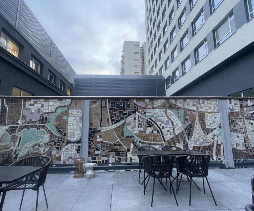 Taras zewnętrzny hotelu Katowice z historyczną mozaiką PRL w tle. Widoczne nowoczesne, czarne stoły i krzesła, obok budynki o ciemnej i jasnej elewacji. Takie architektoniczne perełki modernizmu znajdziesz na Architektura Murator Plus.