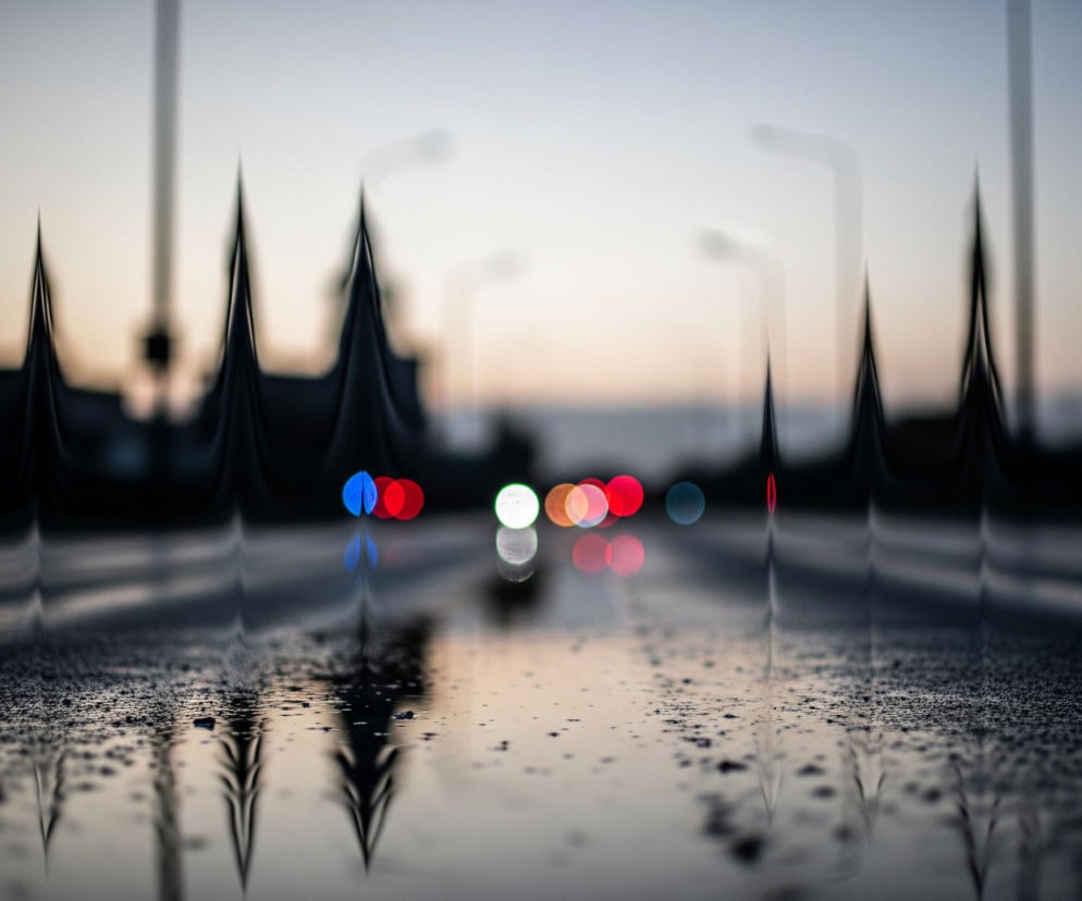 Na pierwszym planie widoczna jest mokra powierzchnia, prawdopodobnie asfalt, pokryta licznymi kroplami wody, która odbija światło. Wzdłuż bocznych krawędzi obrazu, zarówno po lewej, jak i po prawej stronie, znajdują się ciemne, pionowe, spiczaste kształty, tworzące szereg podobnych do gór lub zębów formacji. W centralnej części obrazu, w tle, widoczne są rozmyte, okrągłe światła w różnych kolorach: czerwonym, zielonym, niebieskim i pomarańczowym, które odbijają się w mokrej nawierzchni. Horyzont jest rozmyty i ma jasne, kremowo-różowe zabarwienie, przechodzące w błękit ku górze.